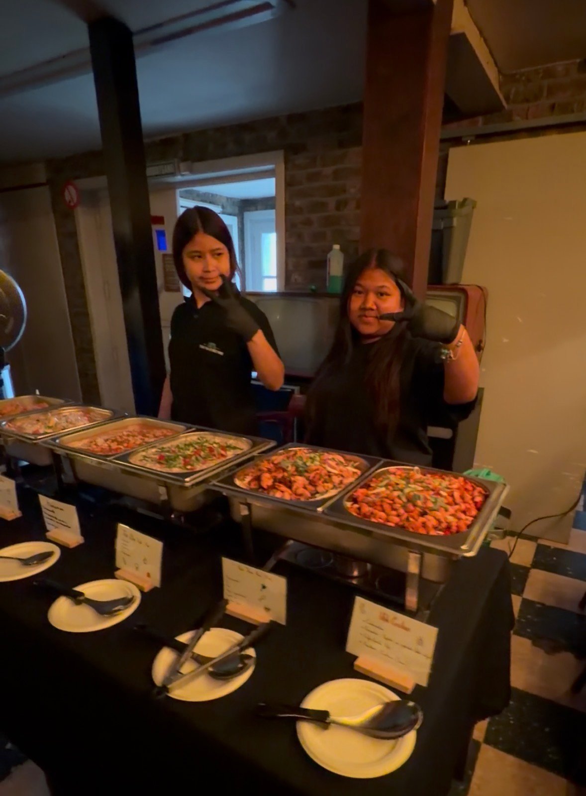 Two restaurant staff members smiling behind a food service counter with multiple metal trays of colorful prepared dishes and empty plates