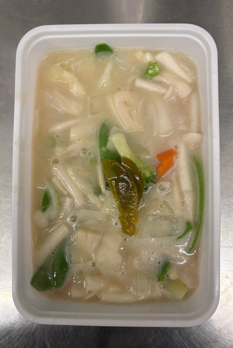 White plastic container of Asian noodle soup with rice noodles, vegetables including green onions and seaweed, and broth