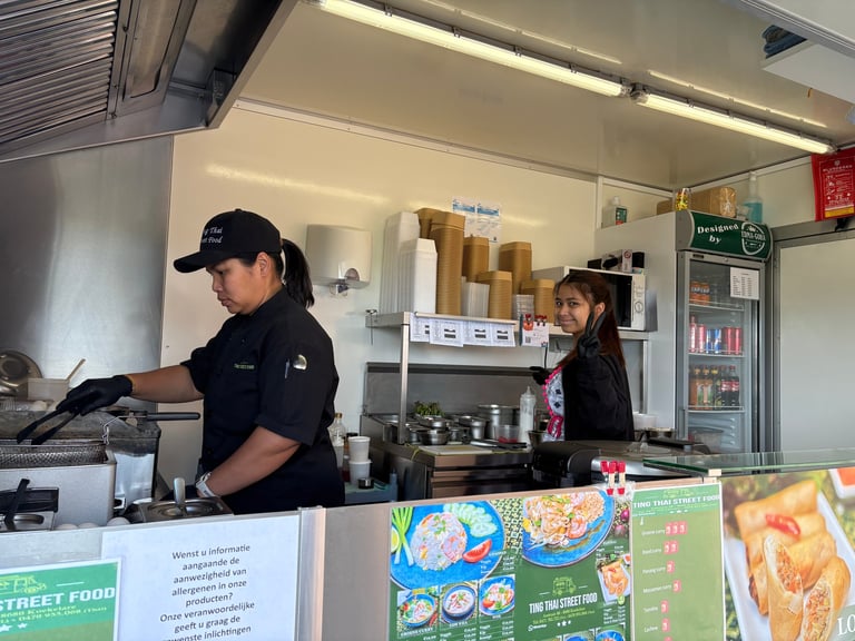 Twee medewerkers werken achter de toonbank in een kleine restaurantkeuken met menuborden en kookuitrusting zichtbaar